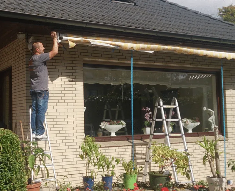 Mann auf einer Leiter, der im Rahmen eines Notdienstes eine Markise über einem Fenster eines Wohnhauses installiert.